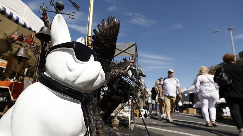 planie znajduje się duża, stylizowana statua buldoga, który nosi czarne okulary przeciwsłoneczne oraz obrożę. Statua, prawdopodobnie wykonana z ceramiki lub podobnego materiału, jest wyeksponowana na stoisku sprzedawcy, który oferuje różnorodne dekoracyjne przedmioty, być może antyki lub kolekcjonerskie ozdoby. Za figurą buldoga widać inne ozdobne przedmioty, takie jak rzeźba orła oraz metalowe elementy. W tle targu można dostrzec kilku ludzi spacerujących po ulicy i przeglądających stragany. Niebo jest jasne, z kilkoma delikatnymi chmurami, co sugeruje ładną pogodę. Cała scena oddaje atmosferę ruchliwego dnia targowego, prawdopodobnie na pchlim targu lub podczas jakiegoś festiwalu, gdzie sprzedawane są różnorodne towary.