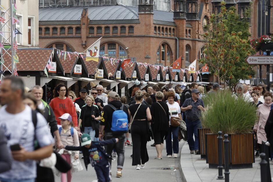 Zdjęcie przedstawia zatłoczoną ulicę podczas Jarmarku Dominikańskiego, odbywającego się w Gdańsku. Wzdłuż ulicy ustawione są rzędy straganów z czerwonymi dachami, na których widoczne są tabliczki z nazwami sprzedawanych produktów. Nad straganami powiewają kolorowe girlandy, dodając festynowego charakteru. Tło zdjęcia stanowi zabytkowa, ceglasta architektura, co dodatkowo podkreśla lokalizację wydarzenia w historycznej części miasta. Tłum ludzi spaceruje po ulicy, niektórzy zatrzymują się przy stoiskach, inni przechodzą dalej. Można dostrzec zarówno rodziny z dziećmi, jak i osoby starsze, co świadczy o różnorodności uczestników tego wydarzenia.