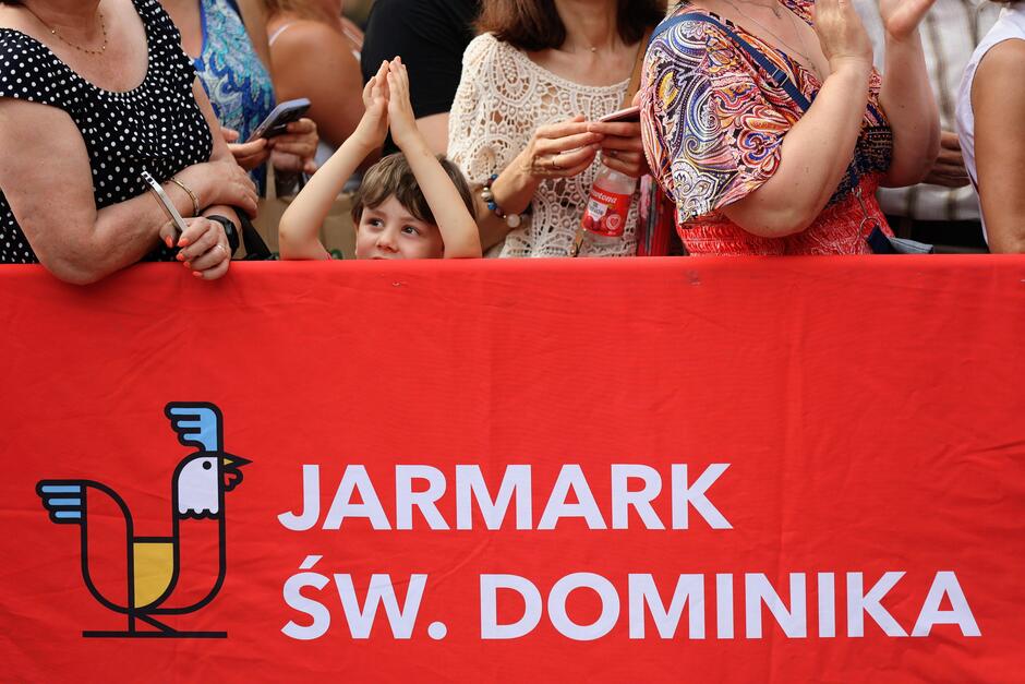 baner z napisem Jarmark św. Dominika, za nim mały chłopiec i gapie