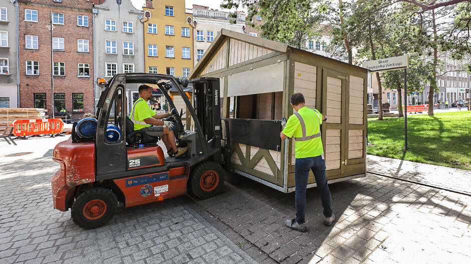 Na zdjęciu widzimy dwóch mężczyzn pracujących przy transporcie drewnianej budki. Jeden z nich obsługuje wózek widłowy marki Nissan, który podnosi konstrukcję, podczas gdy drugi mężczyzna nadzoruje całą operację, stojąc obok budki. Obaj pracownicy mają na sobie jaskrawe, żółte kamizelki odblaskowe. Scena rozgrywa się na miejskim placu, na którym widać brukowaną nawierzchnię i otaczające go budynki. W tle dostrzegamy kamienice, charakterystyczne dla europejskich miast, oraz zielony trawnik z drzewami. Widoczna jest także tabliczka z napisem Skwer Świętopełka Wielkiego,  co sugeruje, że zdjęcie zostało wykonane w Polsce, prawdopodobnie w Gdańsku.