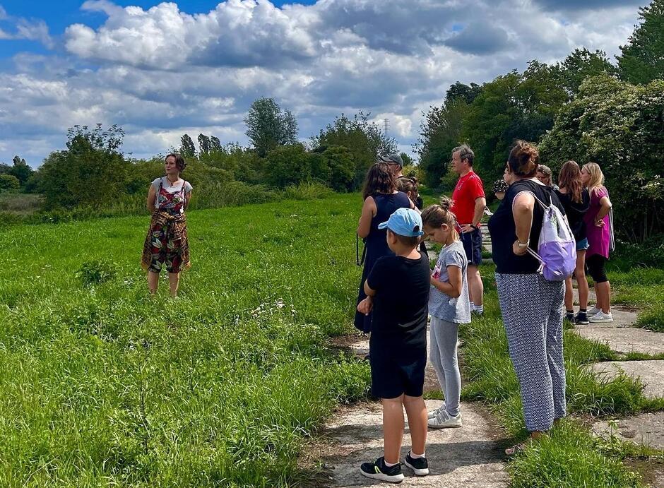 Zdjęcie przedstawia grupę osób, w tym dzieci i dorosłych, uczestniczących w spacerze po zielonym terenie na świeżym powietrzu, słuchających kobiety w kolorowej sukience, która prawdopodobnie pełni rolę przewodnika. W tle widać zalesiony obszar, a nad uczestnikami unosi się niebo pokryte białymi chmurami