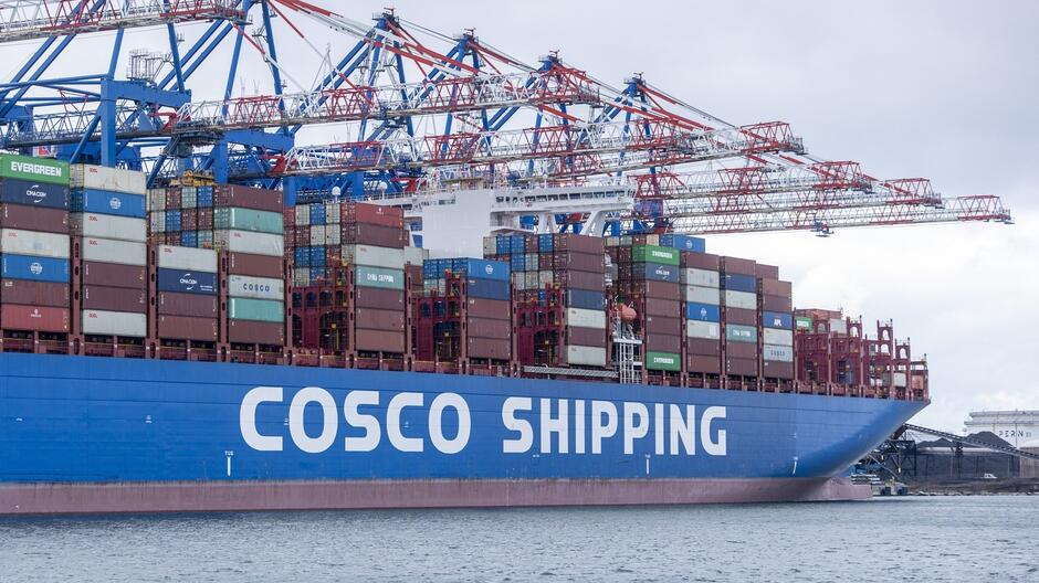 Na zdjęciu widać duży kontenerowiec z logo COSCO SHIPPING  na burcie, który jest zacumowany przy nabrzeżu. Na pokładzie statku znajdują się stosy kolorowych kontenerów różnych firm, ułożone w wysokie rzędy. W tle widać masywne dźwigi portowe, używane do załadunku i rozładunku kontenerów. Niebo jest zachmurzone, a woda w porcie spokojna. Cała scena przedstawia typowy widok z portu morskiego, związanego z transportem towarów na skalę międzynarodową.