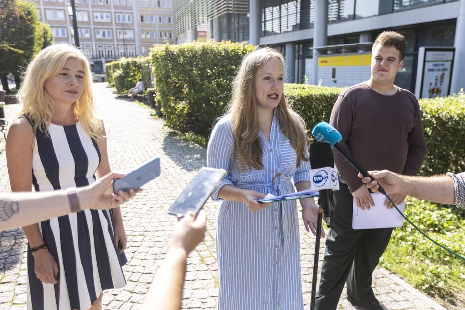 Z lewej strony szczupła kobieta w średnim wieku z długimi blond włosami w sukience w paski obok niej młoda kobieta z długimi jasnymi włosami i z prawej młody chłopak w ciemnej koszulce