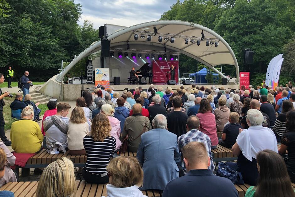 Widzimy od tyłu publiczność zasiadającą w amfiteatrze. W dalszym tle jest scena z artystką