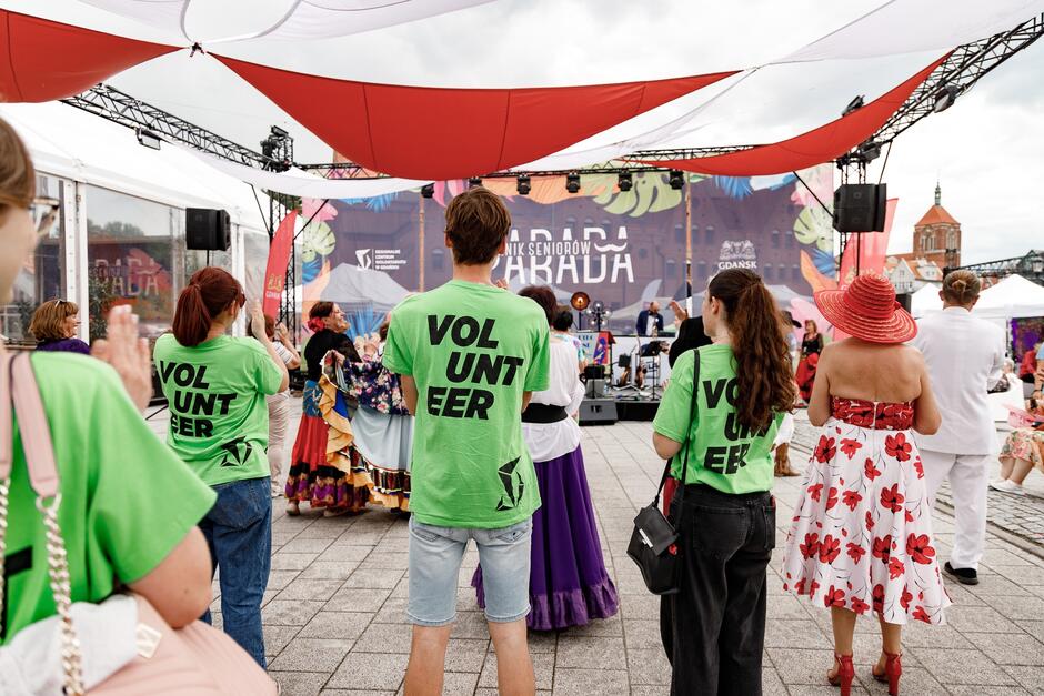 Trzy osoby w zielonych koszulkach z napisem volunteer  stoją tyłem do aparatu i obserwują zabawę seniorów podczas pikniku i parady seniorów w Gdańsku.