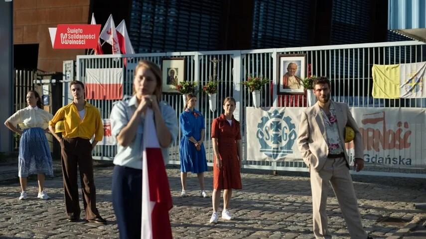 Na zdjęciu znajduje się grupa młodych ludzi ubranych w stroje stylizowane na lata 80., stojących przed bramą ozdobioną flagami i plakatami związanymi z ruchem Solidarności w Polsce. Tło tworzy przemysłowa sceneria, w tle obecność flag i portretów papieża Jana Pawła II oraz symboli Solidarności