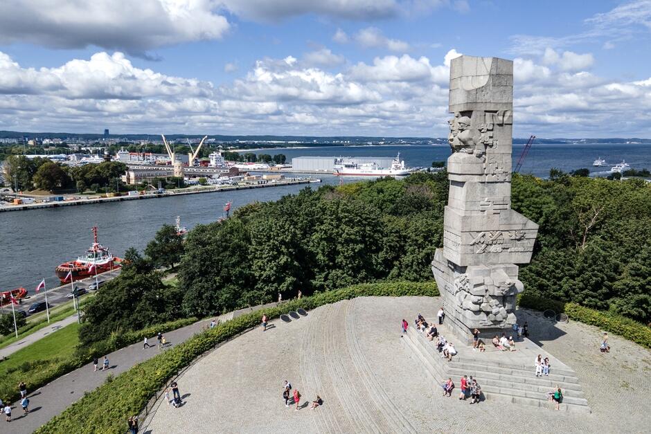 Na zdjęciu widoczny jest Pomnik Obrońców Wybrzeża znajdujący się na Westerplatte w Gdańsku, Polska, upamiętniający polskich żołnierzy, którzy bronili wybrzeża w czasie II wojny światowej. W tle widać port morski, statki oraz krajobraz nadmorski z zielonymi drzewami i rozległym niebem z chmurami