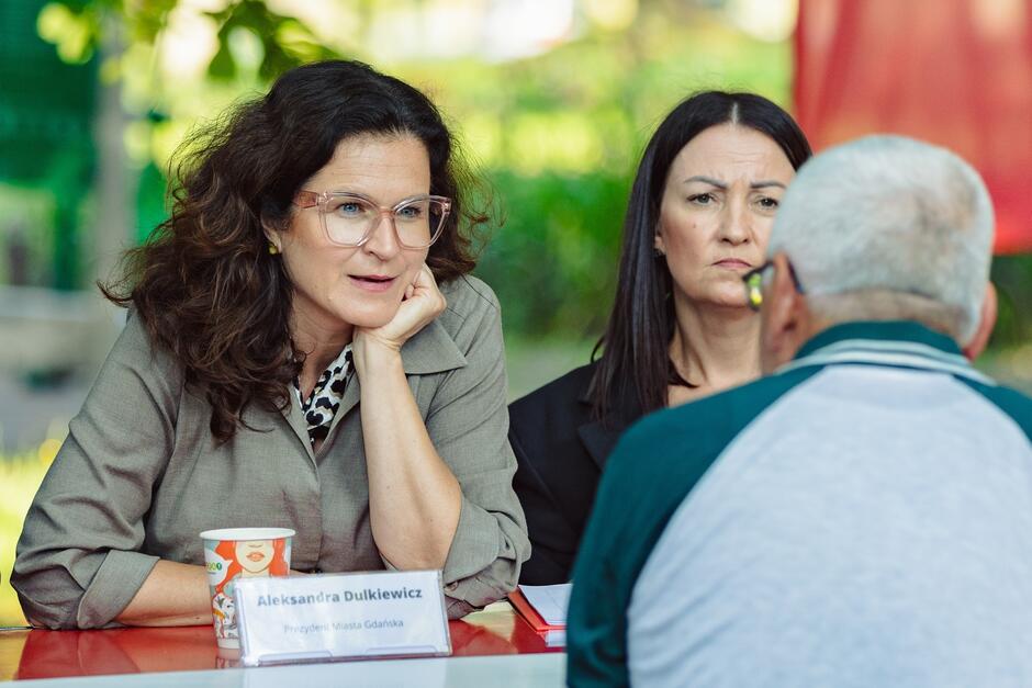 na zdjęciu kobieta w średnim wieku, z długimi ciemnymi włosami i okularami, w szarej sukience, rozmawia z mężczyzną, który odwrócony jest tyłem do fotografującego, widać ich od pasa w górę