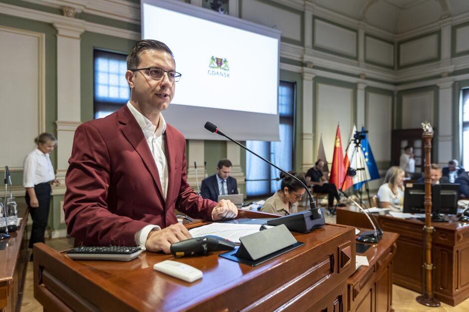 Na zdjęciu znajduje się mężczyzna w bordowej marynarce, który stoi przy mównicy w sali obrad, prawdopodobnie podczas oficjalnego spotkania lub sesji rady miejskiej. Mężczyzna wygląda na skupionego, patrzy przed siebie, trzymając ręce na mównicy. Przed nim znajduje się mikrofon, z którego korzysta, aby przemawiać do zgromadzonych. W tle widać duży ekran, na którym wyświetlone jest logo miasta Gdańsk, co sugeruje, że spotkanie ma miejsce w Gdańsku. Po obu stronach ekranu znajdują się okna z zasłoniętymi roletami. Widać również kilka flag stojących w rogu pomieszczenia, co nadaje miejscu oficjalny charakter. Przy stołach siedzi kilku innych uczestników, zajętych swoimi zadaniami, a niektórzy z nich używają komputerów lub rozmawiają. Atmosfera jest formalna, a wnętrze, podobnie jak na poprzednim zdjęciu, ma klasyczny wystrój z elementami drewnianymi i sztukateriami na ścianach.