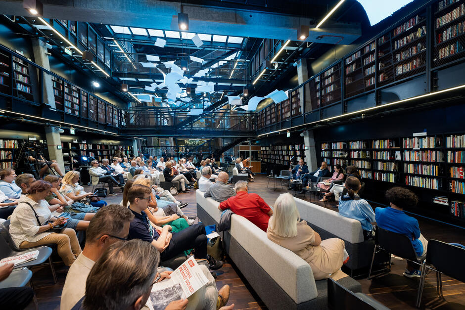 Zdjęcie przedstawia nowoczesne wnętrze biblioteki, w której odbywa się spotkanie lub wykład z udziałem licznej publiczności siedzącej na krzesłach i sofach. W tle widać regały pełne książek, a nad uczestnikami zawieszona jest instalacja artystyczna z białych papierów lub kart