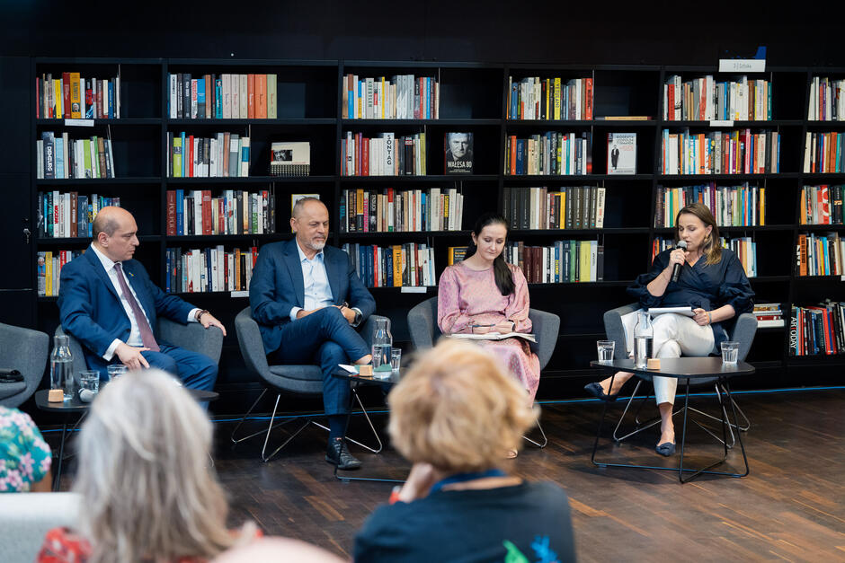 Zdjęcie przedstawia czterech panelistów siedzących na krzesłach przed półkami pełnymi książek podczas dyskusji lub panelu dyskusyjnego. Każdy z nich jest skupiony na swojej roli, a jedna z kobiet trzyma mikrofon i mówi, podczas gdy pozostali słuchają lub mają notatki przed sobą