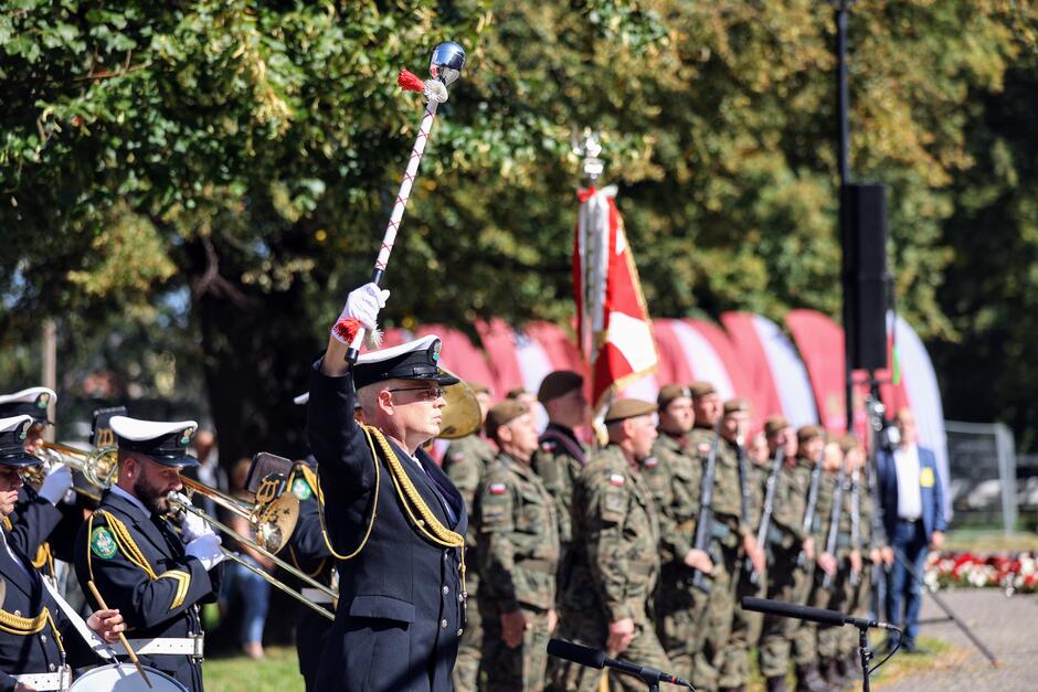 Na zdjęciu widoczna jest parada wojskowa, gdzie na pierwszym planie mężczyzna w mundurze marynarki wojennej unosi buławę, prowadząc orkiestrę wojskową. W tle widać żołnierzy w mundurach polowych oraz czerwono-białe flagi, co sugeruje uroczystość o charakterze patriotycznym.