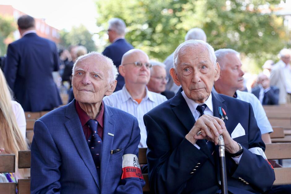 Na zdjęciu widzimy dwóch starszych mężczyzn siedzących obok siebie podczas uroczystości, ubranych w eleganckie garnitury; jeden z nich nosi opaskę z napisem Szare Szeregi . W tle widać innych uczestników uroczystości