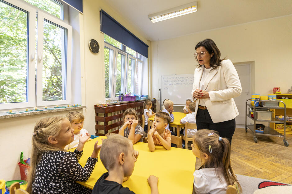 elegancko ubrana kobieta w okularach rozmawia z siedzącymi przy stole małymi dziećmi. w tle wnętrze szkolnej klasy, na ścianach widać kolorowe plakaty i rysunki. za oknami dużo zieleni