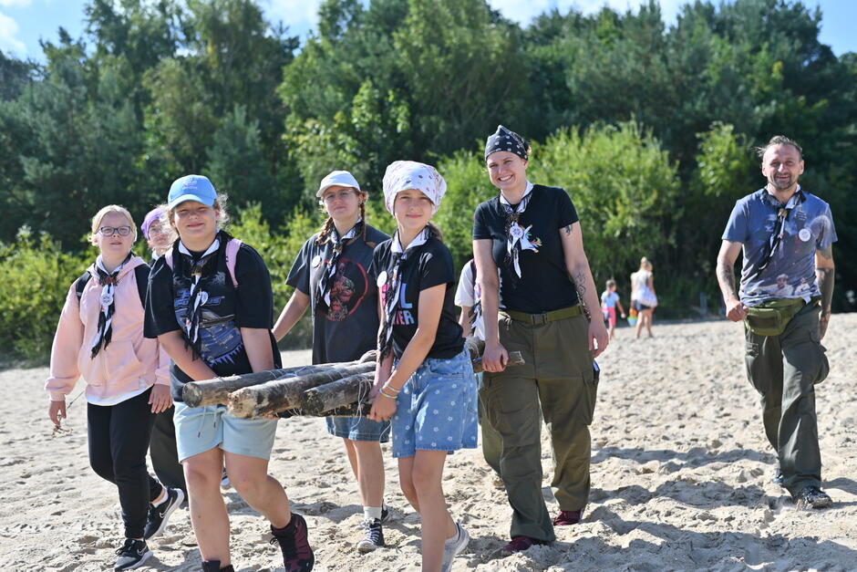 Kilkoro młodych ludzi niesie drwa na plaży, w tle las. 