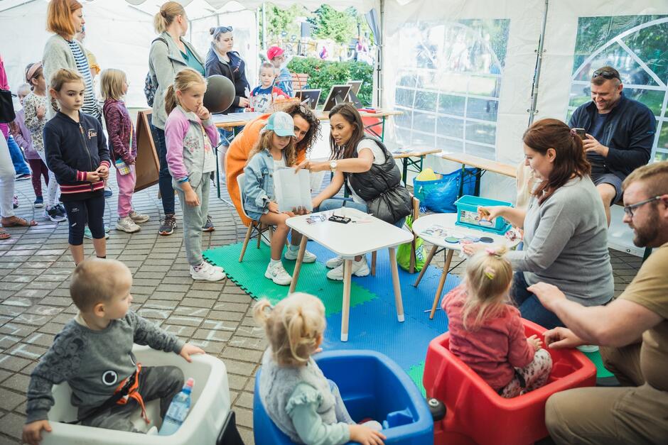 na zdjęciu duży biały namiot, a pod nim sporo dzieci, niektóre stoją, kilka siedzi w plastikowych dużych siedziskach, jest też kilku dorosłych, którzy im się przyglądają