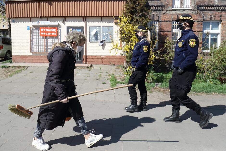 Ulica którą przechodzi dwoje strażników miejskich w maseczkach sanitarnych na twarzy. Idą z prawej strony zdjęcia w lewo. W przeciwnik kierunku idzie starsza pani z miotłą i kubełkiem, również ma maseczkę. W tle stara zabudowa, w niezbyt dobrym stanie