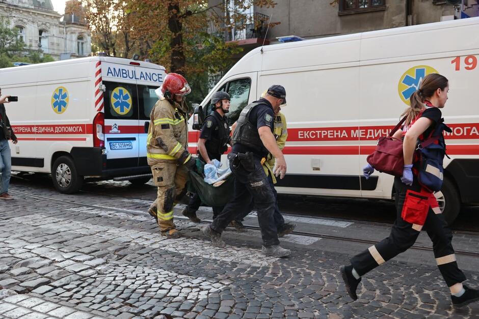 Troje ratowników niesie na kocu jakąś osobę, którą widać tylko częściowo. W tle ambulanse pogotowia ratunkowego