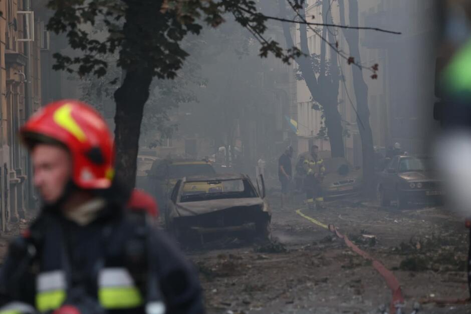 Ulica zasnuta dymem pożaru, widoczne zniszczenia. Na pierwszym planie, nieco niewyraźny stoi strażak w pełnym ekwipunku ratowniczym