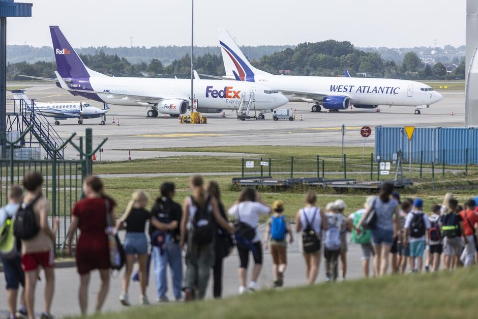 Zdjęcie przedstawia grupę dzieci spacerujących w kierunku lotniska, na którym w tle widać stojące samoloty, w tym maszyny oznaczone logotypami FedEx oraz West Atlantic. Dzieci są ubrane w letnie stroje i plecaki, co sugeruje, że mogą uczestniczyć w wycieczce lub pikniku na terenie lotniska.