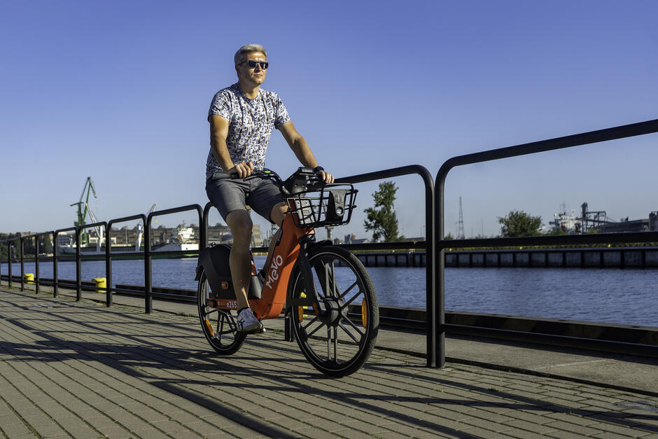 Zdjęcie przedstawia mężczyznę jadącego na rowerze miejskim marki Mevo  wzdłuż nabrzeża. Ubrany w letni strój, z krótkimi spodenkami i koszulką, mężczyzna ma na sobie okulary przeciwsłoneczne, a w tle widoczna jest przystań z żurawiami portowymi oraz spokojna woda, co sugeruje, że zdjęcie zostało wykonane w słoneczny dzień nad morzem lub rzeką.