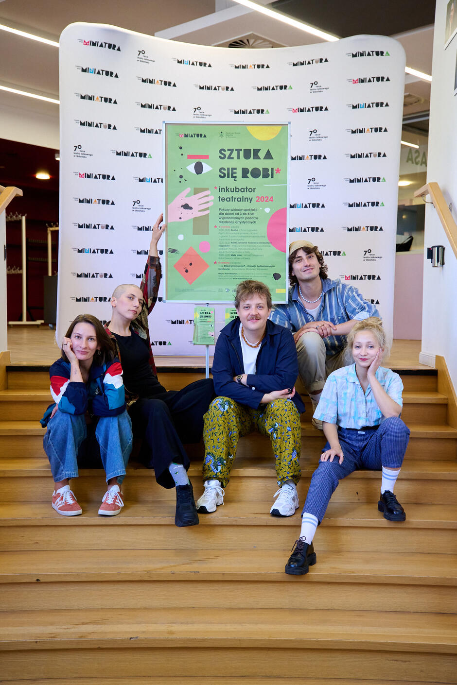 Grupa osób siedzi na drewnianych schodach wewnątrz budynku teatru. Za nimi stoi ścianka a na niej zielony plakat z opisem wydarzenia