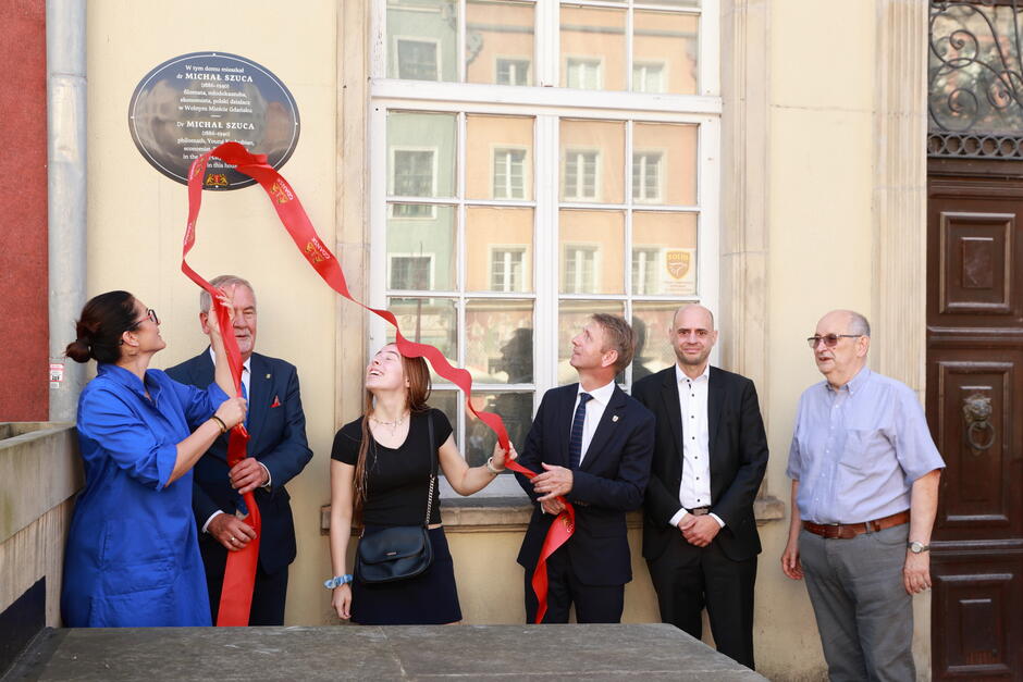 Na zdjęciu widzimy grupę sześciu osób stojących przed budynkiem, uczestniczących w ceremonii odsłonięcia tablicy pamiątkowej. Tablica dedykowana jest Michałowi Szuca, a dwie osoby trzymają wstęgę, która właśnie została przecięta, co sugeruje uroczyste upamiętnienie jego osoby.