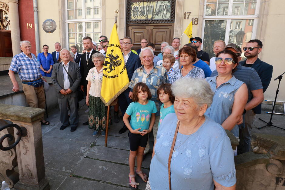 Na zdjęciu znajduje się duża grupa ludzi, w tym starsze osoby, dzieci oraz dorośli, zgromadzona przed budynkiem z numerami 17 i 18, gdzie widoczna jest pamiątkowa tablica poświęcona Michałowi Szuca. Jedna z osób trzyma sztandar z herbem przedstawiającym czarnego gryfa, co sugeruje, że uroczystość ma znaczenie patriotyczne lub związane z lokalną historią.