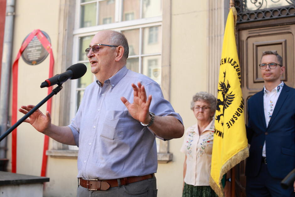 Na zdjęciu starszy mężczyzna w okularach przemawia do mikrofonu podczas uroczystości, trzymając ręce w geście podkreślającym swoje słowa. W tle widoczna jest tablica pamiątkowa, ozdobiona wstęgą, oraz osoby towarzyszące, w tym ktoś trzymający sztandar z kaszubskim gryfem.