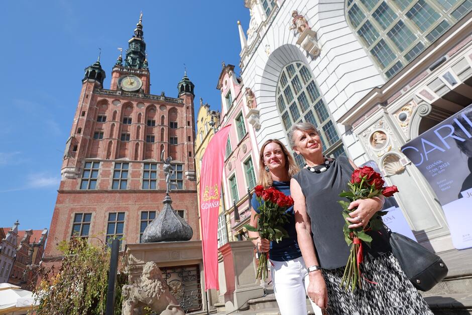 Na zdjęciu widać dwie uśmiechnięte kobiety trzymające bukiety czerwonych róż, stojące przed zabytkowymi budynkami w Gdańsku, w tym charakterystyczną Wieżą Ratusza Głównego Miasta. W tle widoczny jest transparent z napisem Gdańsk , a także elementy architektury renesansowej, które nadają scenie historyczny charakter.