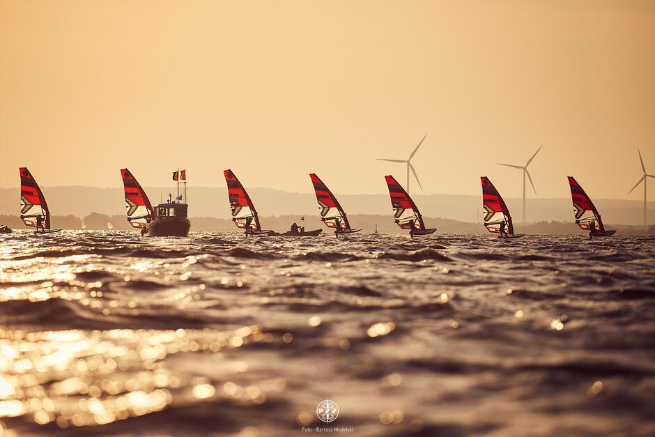 Na zdjęciu widać grupę windsurferów na wodzie z czerwono-czarnymi żaglami, sunących w stronę motorówki, która znajduje się pośrodku kadru. W tle widać farmę wiatrową z turbinami na horyzoncie, a wszystko to jest oświetlone ciepłym światłem zachodzącego słońca.