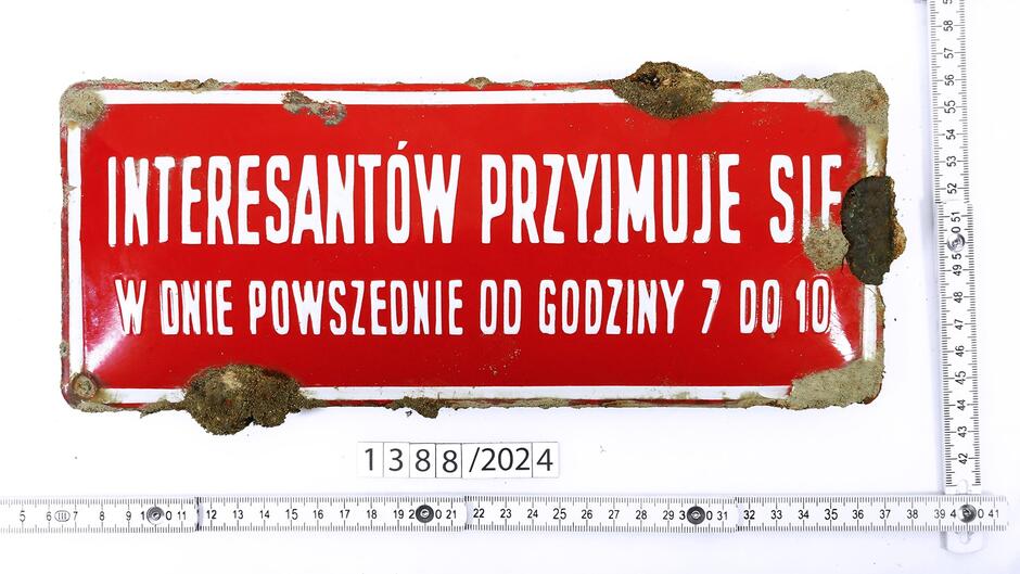 Na zdjęciu widoczna jest stara metalowa tabliczka o czerwonym tle z białym napisem w języku polskim, która nosi ślady rdzy oraz zniszczeń, zwłaszcza w rogach. Napis głosi: INTERESANTÓW PRZYJMUJE SIĘ W DNIE POWSZECHNIE OD GODZINY 7 DO 10 , co oznacza, że interesanci są przyjmowani w dni powszednie od godziny 7 do 10. Wokół tabliczki umieszczono linijki pomiarowe wskazujące jej wymiary. Na dole zdjęcia widnieje dodatkowy numer identyfikacyjny:  1388/2024 , prawdopodobnie odnoszący się do procesu katalogowania bądź identyfikacji tego obiektu.