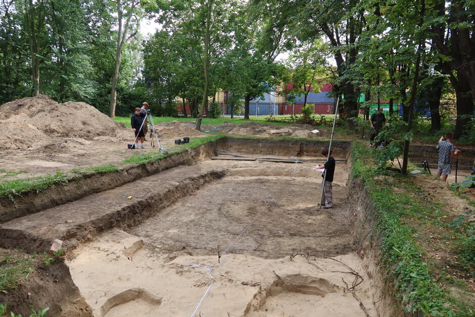 Na zdjęciu przedstawiono teren wykopalisk archeologicznych znajdujący się w otoczeniu drzew i zieleni. Na pierwszym planie widoczny jest duży wykop podzielony na sekcje, z odsłoniętą warstwą gleby i piasku. Wzdłuż krawędzi wykopu widoczne są różne poziomy warstw ziemi. W różnych miejscach widać osoby prowadzące prace pomiarowe i dokumentacyjne. Po lewej stronie dwie osoby korzystają z urządzeń geodezyjnych, a po prawej stronie inna osoba trzyma pionową tyczkę pomiarową i notuje dane. Prace odbywają się na świeżym powietrzu, w otoczeniu drzew, co wskazuje na to, że wykopaliska mają miejsce w parku lub zalesionym terenie. W tle widoczne są budynki przemysłowe lub kontenery za ogrodzeniem. Fotografia dokumentuje zaawansowane prace archeologiczne, w których wykonywane są pomiary, zapewniające dokładne zarejestrowanie lokalizacji znalezisk.