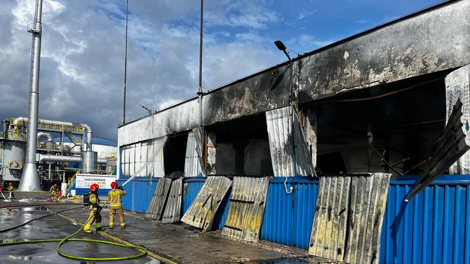 Na zdjęciu widoczny jest budynek przemysłowy, który uległ poważnemu uszkodzeniu na skutek pożaru. Elewacja budynku jest silnie nadpalona i osmolona, a metalowe panele ścienne są wygięte i oderwane od konstrukcji, prawdopodobnie w wyniku wysokiej temperatury. Części ścian są zniszczone, odsłaniając wnętrze budynku. Na pierwszym planie widoczni są strażacy w pełnym wyposażeniu, ubrani w żółte mundury ochronne i czerwone hełmy. Jeden z nich trzyma wąż strażacki i polewa wodą ścianę budynku, zapewne w celu dogaszania pozostałości po pożarze. W tle po lewej stronie znajdują się inne elementy infrastruktury przemysłowej, w tym rurociągi i zbiorniki, sugerujące, że miejsce to mogło być częścią zakładu przemysłowego lub fabryki. Tło jest częściowo zasnute chmurami, jednak na zdjęciu widać też fragmenty niebieskiego nieba, co sugeruje, że pogoda była zmienna. Fotografia ukazuje akcję straży pożarnej i skutki pożaru w zakładzie przemysłowym.