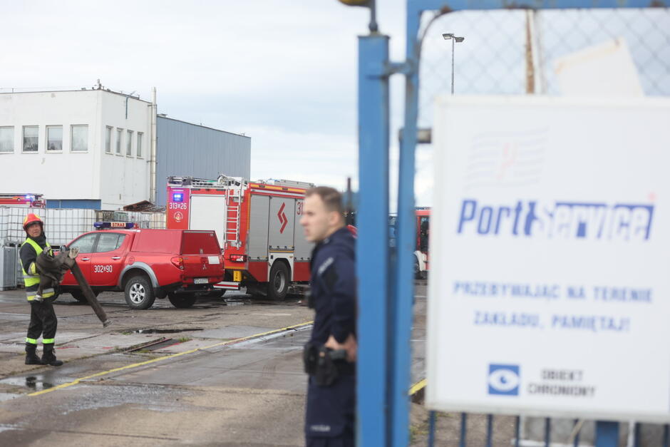 Na zdjęciu widoczna jest akcja ratunkowa prowadzona przez straż pożarną na terenie przemysłowym firmy Port Service. W tle widać pojazdy straży pożarnej, w tym czerwony samochód operacyjny oraz wóz strażacki z drabiną, oznaczony numerami 313[26] . Strażacy są w trakcie zwijania sprzętu po akcji gaśniczej lub ratunkowej – jeden z nich trzyma wąż strażacki. Z przodu po prawej stronie znajduje się tabliczka z nazwą firmy Port Service i informacją o tym, że teren zakładu jest obiektem chronionym. Przed tabliczką stoi funkcjonariusz policji, co wskazuje na współpracę służb w zabezpieczeniu miejsca zdarzenia. W tle widać także budynki przemysłowe oraz metalowe bariery, co sugeruje, że akcja miała miejsce na zamkniętym terenie zakładu przemysłowego. Atmosfera zdjęcia sugeruje, że akcja ratunkowa jest już w końcowej fazie, a strażacy i służby porządkowe zajmują się porządkowaniem sprzętu.