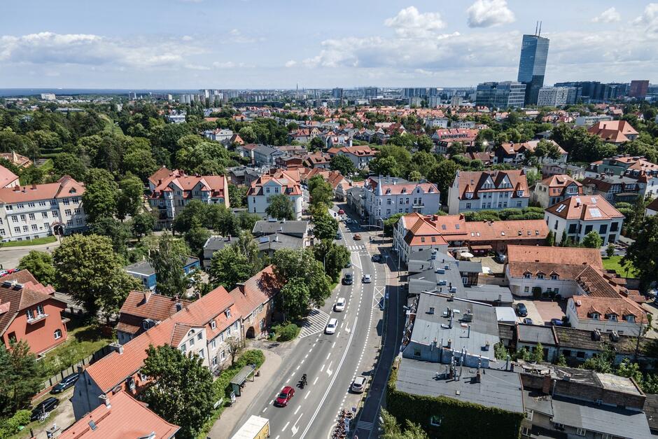 zdjęcie z drona, widać na pierwszym planie dużo domów jednorodzinnych otoczonych zielonymi dużymi drzewami, w tle kilka wysokich biurowców