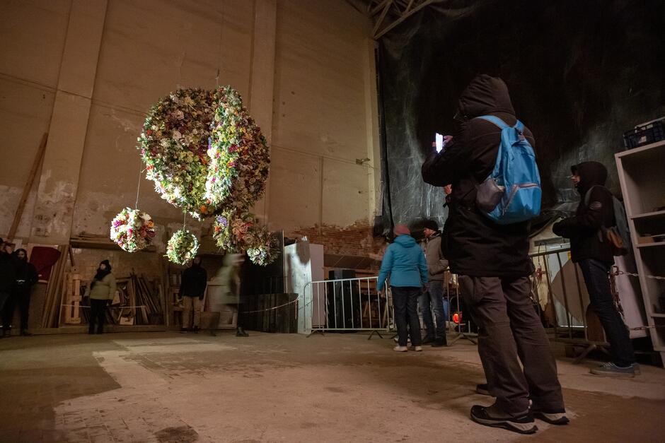 Na zdjęciu widzimy instalację artystyczną, prawdopodobnie z jakiegoś festiwalu sztuki, który odbywa się w surowym, przemysłowym wnętrzu. W centrum kadru wiszą różne obiekty pokryte sztucznymi lub suszonymi kwiatami, które tworzą duże kuliste kształty. Instalacja jest podwieszona pod sufitem i oświetlona z boku, co nadaje całości ciepłego, przyjemnego efektu. Wokół instalacji stoi grupa osób, z których niektóre obserwują dzieło, a inne robią zdjęcia. Ludzie są ubrani w ciepłe kurtki, co sugeruje, że jest zimno, prawdopodobnie festiwal odbywa się w chłodnej porze roku lub w nieogrzewanym budynku. Tło jest surowe, z widocznymi ścianami pokrytymi łuszczącą się farbą oraz z elementami budowlanymi i drewnem ustawionym w kącie. Atmosfera wydaje się spokojna, a ludzie uważnie przyglądają się instalacji.