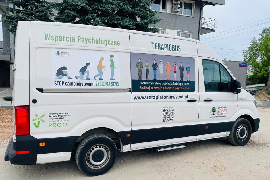 Zdjęcie przedstawia biały samochód oznaczony jako Terapiobus , który oferuje wsparcie psychologiczne. Na bocznej ścianie pojazdu znajduje się plakat promujący zapobieganie samobójstwom oraz hasła zachęcające do dbania o zdrowie psychiczne, z informacją o stronie internetowej kampanii  terapiatoniewstyd.pl .