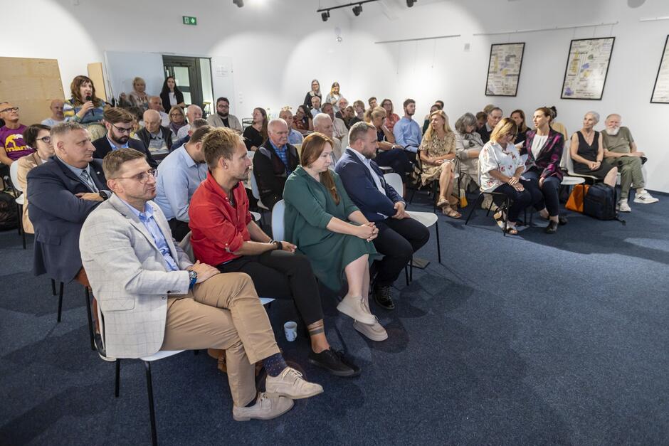 Kilkadziesiąt osób siedzi na krzesłach w sali konferencyjnej. jedna kobieta stoi i mówi do mikrofonu.