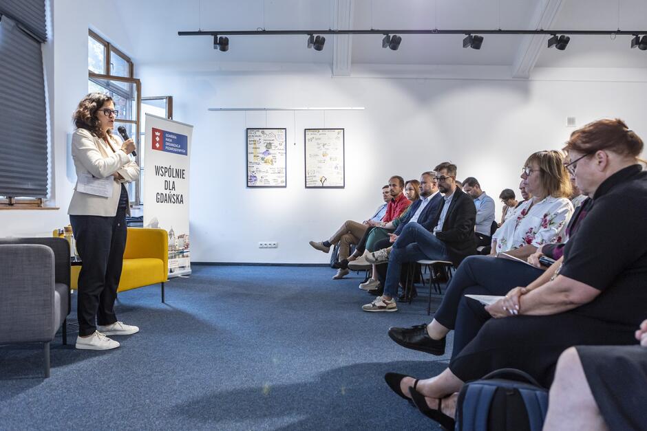 Kobieta stoi i mówi do mikrofonu w sali konferencyjnej, zwrócona przodem do ludzi siedzących na krzesłach. 