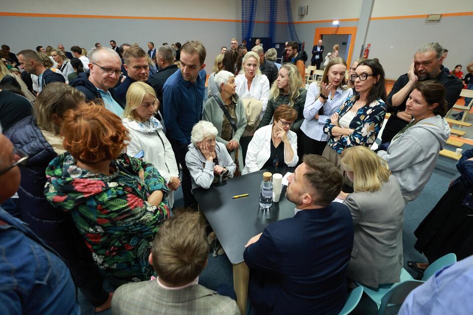 na zdjęciu kilkanaście osób w średnim wieku stoi wokół niewielkiego stolika przy którym siedzi mężczyzna w garniturze i kilka innych osób zwyczajnie ubranych, w tle widać tłum ludzi stojących w dużym pomieszczeniu