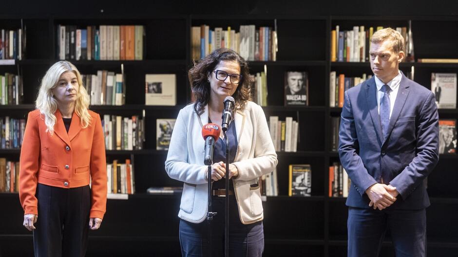 Na zdjęciu widać troje ludzi stojących przed półkami z książkami, w otoczeniu przypominającym bibliotekę lub księgarnię. Kobieta w środku, ubrana w białą marynarkę i okulary, przemawia do mikrofonu z logiem TOKS , trzymając ręce złożone przed sobą. Po jej lewej stronie stoi kobieta w czerwonej marynarce, z długimi, blond włosami, patrząca poważnie. Po prawej stronie stoi mężczyzna w granatowym garniturze i krawacie, z rękami splecionymi z przodu. W tle widoczne są półki wypełnione książkami, z niektórymi tytułami widocznymi na okładkach.