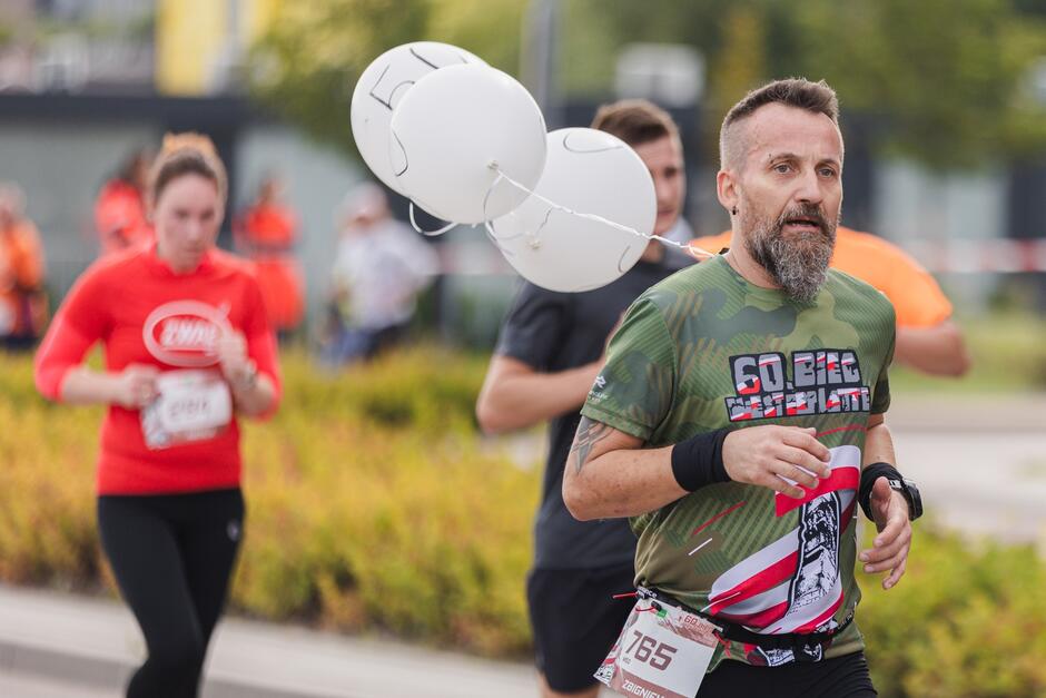 Na zdjęciu widać mężczyznę biorącego udział w biegu, ubrany jest w zieloną koszulkę z napisem 60 BIG  i przypiętym numerem startowym. Za nim biegną inni uczestnicy, a do mężczyzny są przyczepione białe balony z narysowanymi cyframi.