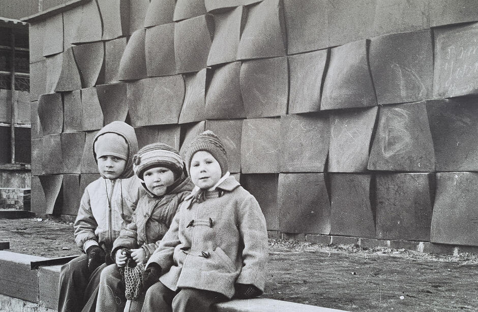 Zdjęcie czarnobiałe z 1977 roku. Na ławce siedzi trójka dzieci w ciepłych ubraniach i czapkach. Za nimi płaskorzeźba plenerowa.