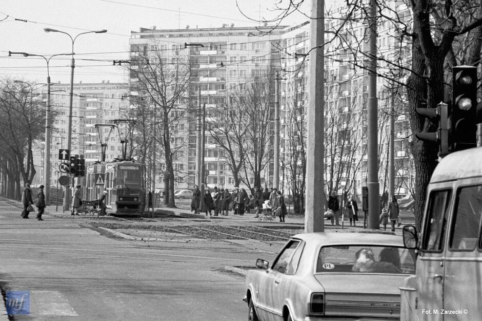 Archiwalne zdjęcie z 1983 roku, czarnobiałe. Ulica na osiedlu mieszkaniowym, która jadą samochody oraz tramwaje, w tle bloki mieszkalne.