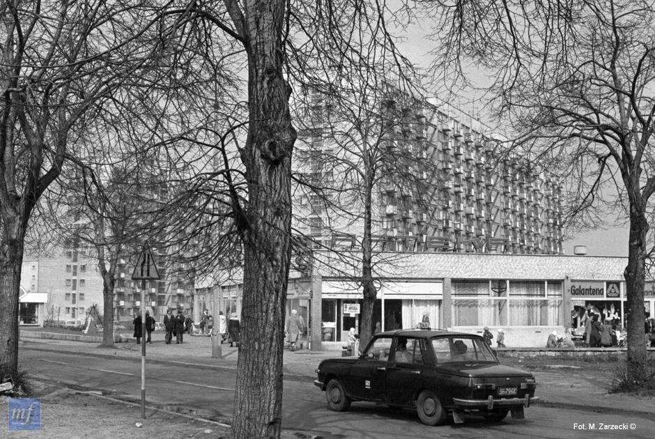 Archiwalne zdjęcie czarnobiałe z 1983 roku. Na pierwszym planie drzewa, między nimi jedzie ulicą samochód wartburg, w tle pawilony sklepowe i bloki mieszkalne.