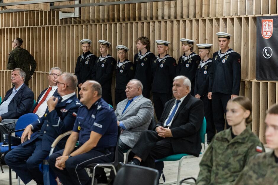 na pierwszym palnie widać kilkunastu starszych panów, w marynarkach bądź mundurach policyjnych, siedzą na krzesłach, za nimi przy ścianie stoi kilku chłopaków w strojach marynarskich