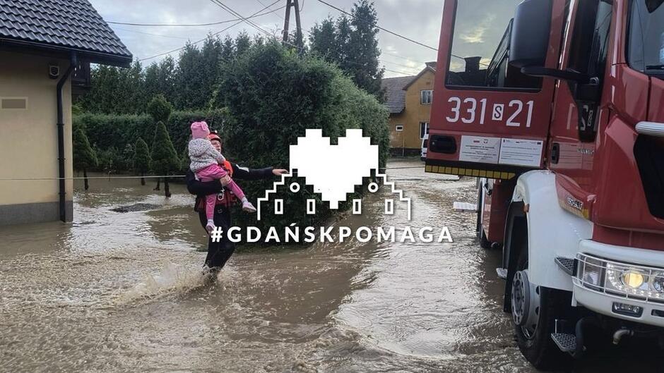 Zdjęcie pokazuje samochód strażacki, który podjeżdża do domu zalanego przez powódź. Na tle zdjęcia jest napis: Gdańsk Pomaga Powodzianom