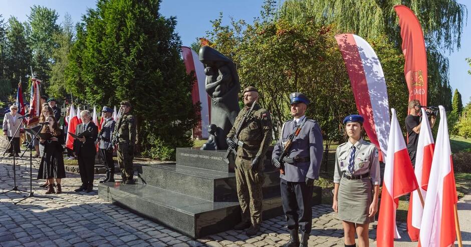 Harcerze i wojskowi przy pomniku pochylonej kobiety z dzieckiem, obok flagi narodowe