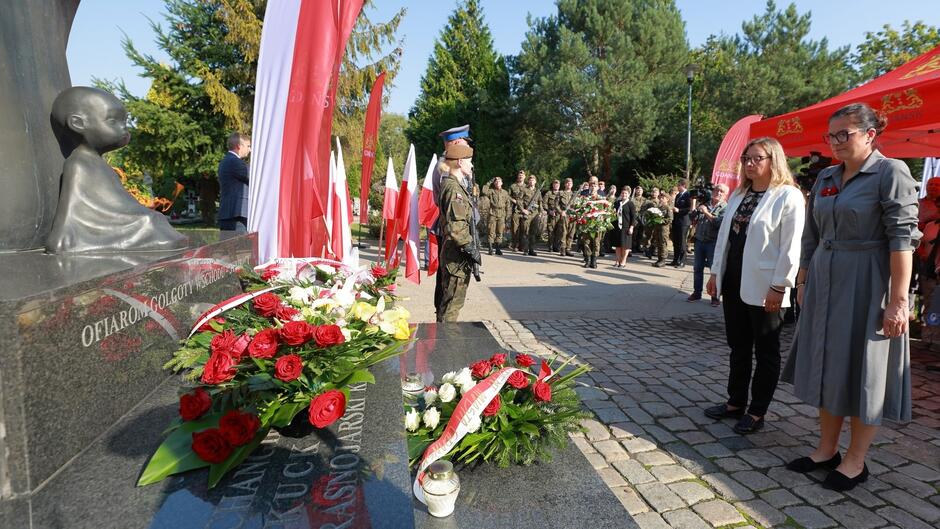 Na zdjęciu widoczna jest uroczystość patriotyczna lub upamiętniająca, odbywająca się w plenerze, prawdopodobnie przy pomniku. Na pierwszym planie znajduje się pomnik z rzeźbą małego dziecka i napisem „OFIAROM GOLGOTY WSCHODU”, symbolizujący hołd dla ofiar. Na pomniku złożone są kwiaty, głównie w kolorach białym i czerwonym, co nawiązuje do barw narodowych Polski. W tle widać grupę osób, w tym żołnierzy w umundurowaniu oraz osoby w cywilnych strojach. Obecność flag oraz oficjalny charakter ustawienia wskazują na uroczystość państwową lub lokalną związaną z ważnym wydarzeniem historycznym. Dwie kobiety w centrum zdjęcia stoją w pozycji honorowej, co podkreśla powagę chwili i szacunek dla upamiętnianych osób. Otoczenie, z drzewami i budynkami, sugeruje, że ceremonia odbywa się na świeżym powietrzu, prawdopodobnie w miejscu pamięci.